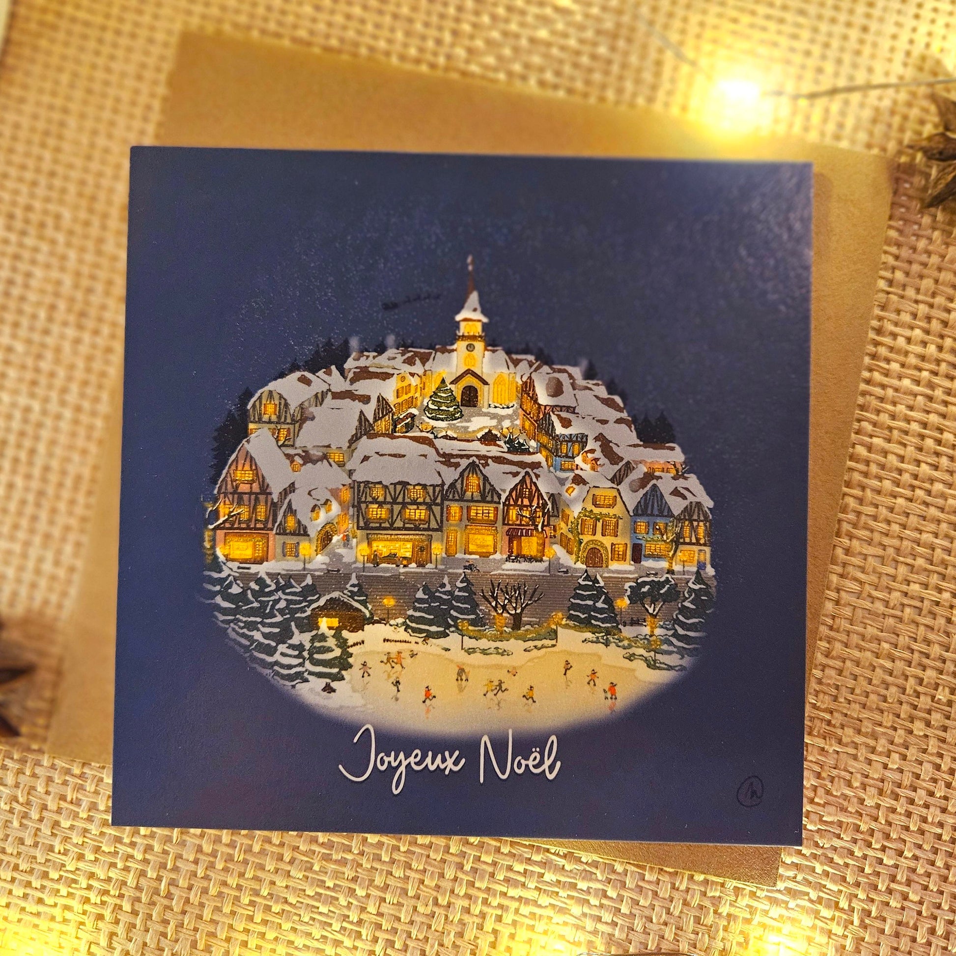 Carte de Noël représentant un village enneigé pendant la période de Noël avec une patinoire en premier plan